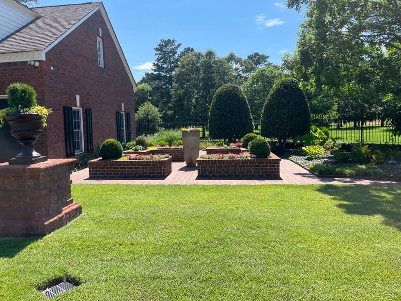 Lush garden with brick planters, manicured shrubs, and vibrant flowers beside a brick home.