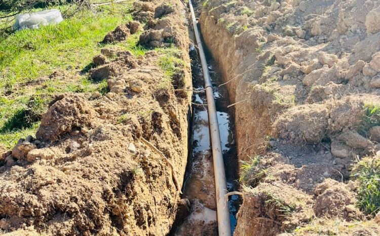 A sizable pipe lies on the ground amidst a wide, open field, surrounded by green grass and distant trees.